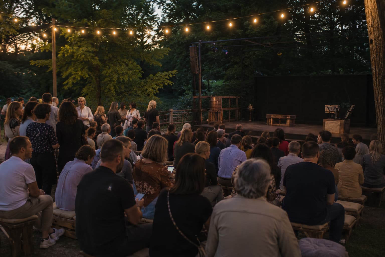 Pubblico raccolto all’aperto prima dell’inizio di uno spettacolo teatrale serale, con palco già allestito e persone sedute o in piedi in attesa.